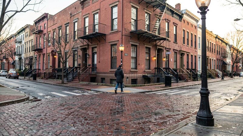 A Baltimore corner at dusk: a terrace of red brick row houses with iron balconies, wet cobblestones catching the light of a cast-iron street lamp, and one figure crossing on the kerb paint.
