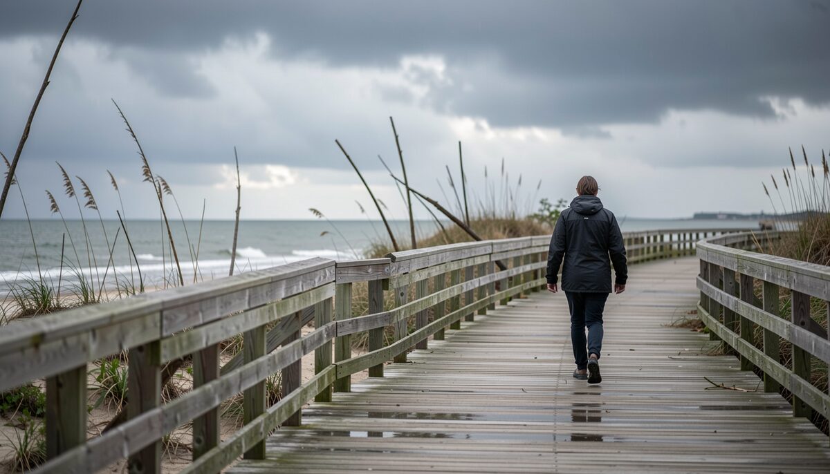 A coastal boardwalk after a storm, bent railings and standing water along the planking, dark cloud cover low over the water and a single figure in a rain jacket walking away.